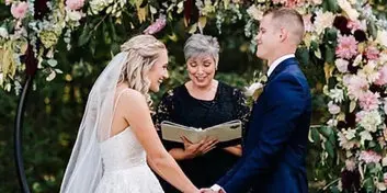 A wedding officiant in a black dress stands between a bride and groom, holding a book and officiating their ceremony under a floral arch.