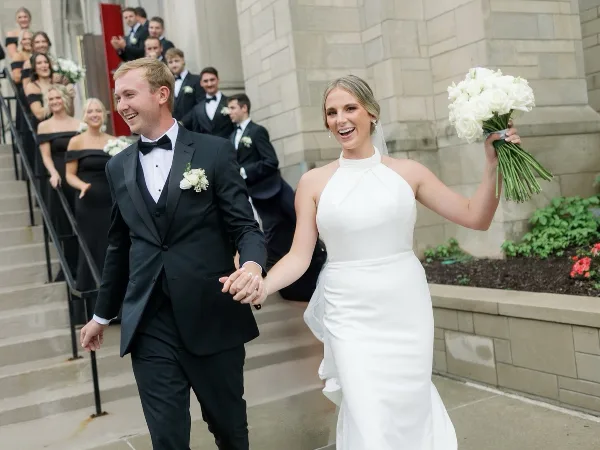 A bride and groom holding hands and smiling as they walk down the steps of a stone building, with wedding party members visible on the stairs behind them.