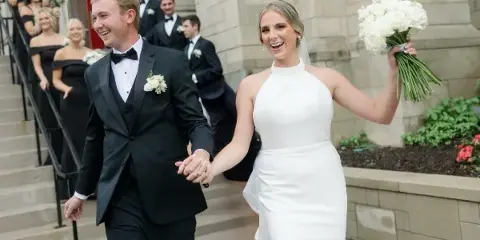 A bride and groom holding hands and smiling as they walk down the steps of a stone building, with wedding party members visible on the stairs behind them.