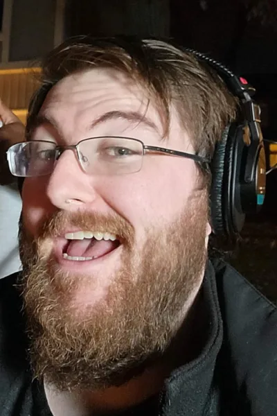 A bearded man wearing glasses and professional over-ear headphones, smiling.