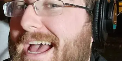 A bearded man wearing glasses and professional over-ear headphones, smiling.
