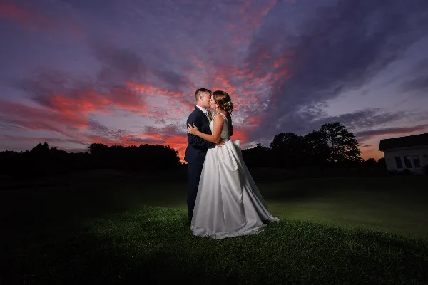 A bride and groom embracing on a grassy field under a dramatic sunset sky.