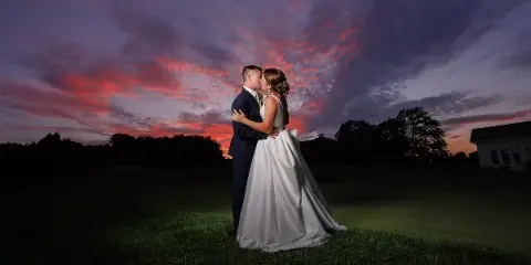 A bride and groom embracing on a grassy field under a dramatic sunset sky.