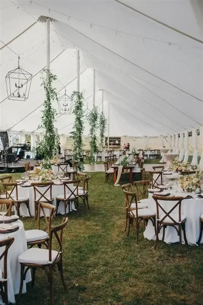 Interior view of a white wedding tent with round tables, wooden chairs, and hanging geometric lanterns.