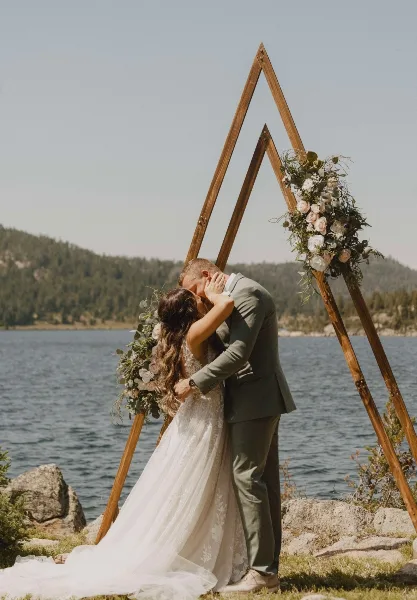 A bride and groom kissing under a wooden A-frame arch decorated with flowers, set against a lake and mountain backdrop.