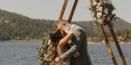 A bride and groom kissing under a wooden A-frame arch decorated with flowers, set against a lake and mountain backdrop.