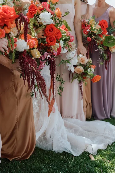 Close-up of a vibrant bridal bouquet and bridesmaids' bouquets featuring red roses, orange blooms, and trailing greenery held by the wedding party.
