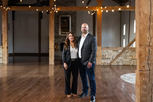 A man and a woman standing in the center of a large, renovated barn interior with exposed wooden beams, string lights, and a stone fireplace.