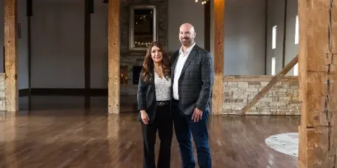 A man and a woman standing in the center of a large, renovated barn interior with exposed wooden beams, string lights, and a stone fireplace.