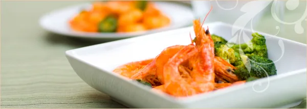 A close-up of a white square plate containing cooked shrimp and broccoli, with another plate of food visible in the background.