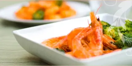 A close-up of a white square plate containing cooked shrimp and broccoli, with another plate of food visible in the background.
