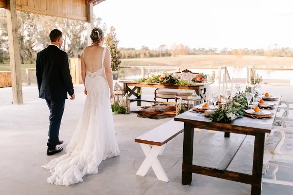 A bride and groom standing in a rustic barn wedding venue with long wooden farm tables and outdoor scenery.
