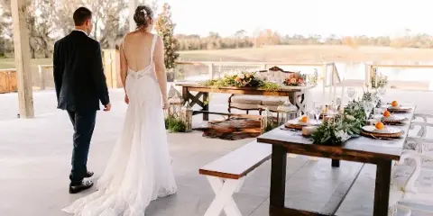 A bride and groom standing in a rustic barn wedding venue with long wooden farm tables and outdoor scenery.