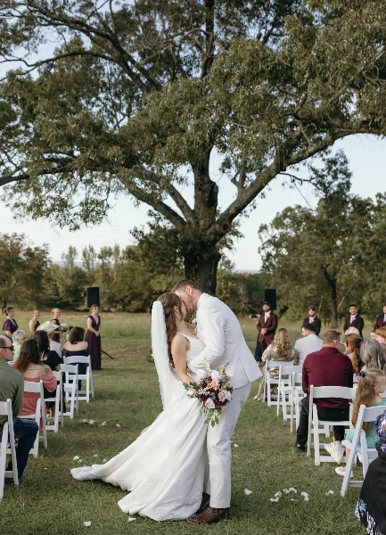 A bride and groom sharing a kiss during an outdoor wedding ceremony under a large tree with guests seated in white chairs.