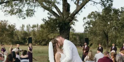 A bride and groom sharing a kiss during an outdoor wedding ceremony under a large tree with guests seated in white chairs.