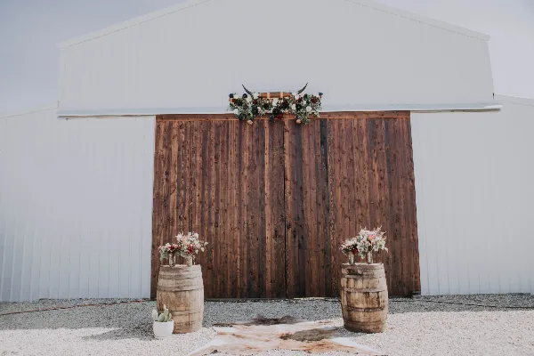 White barn exterior with wooden doors decorated with a floral arch and barrel arrangements.
