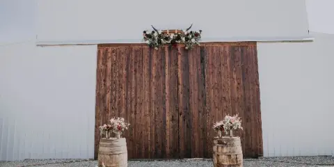White barn exterior with wooden doors decorated with a floral arch and barrel arrangements.