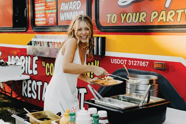 A smiling woman in a white dress serving food from a mobile catering truck with chafing dishes and condiments.