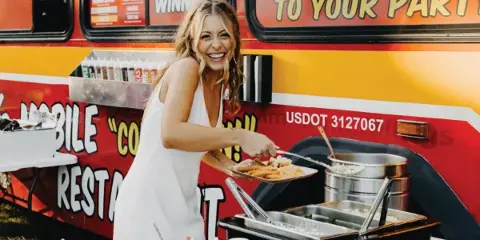 A smiling woman in a white dress serving food from a mobile catering truck with chafing dishes and condiments.