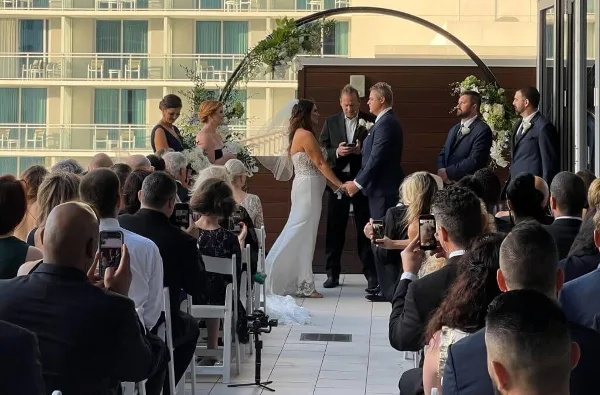 A wedding ceremony taking place on an outdoor terrace with a bride and groom at an altar, officiant, and seated guests.