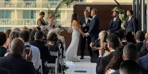 A wedding ceremony taking place on an outdoor terrace with a bride and groom at an altar, officiant, and seated guests.