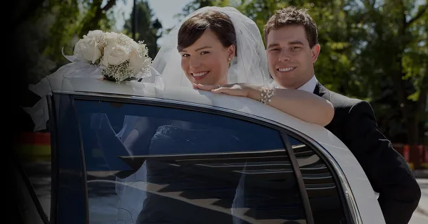 A bride and groom smiling while leaning on a white car decorated with a white rose bouquet.