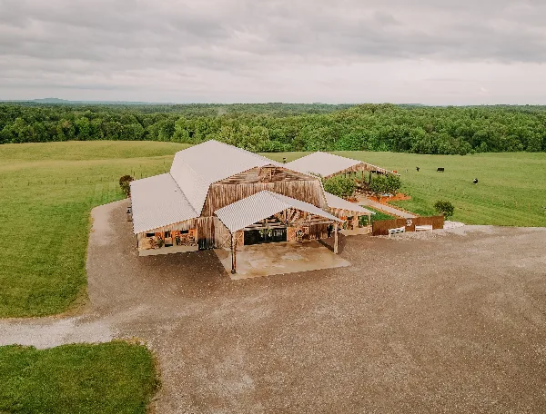 Aerial view of a rustic barn wedding venue with metal roofs, outdoor event spaces, and surrounding green farm fields.
