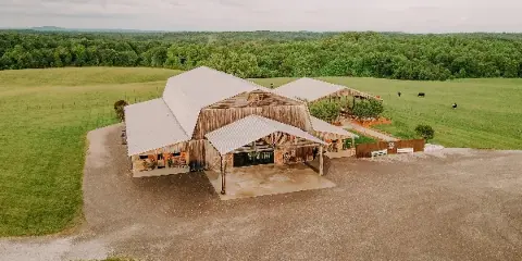 Aerial view of a rustic barn wedding venue with metal roofs, outdoor event spaces, and surrounding green farm fields.