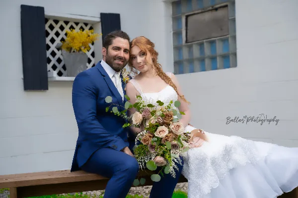 A bride and groom posing for a wedding portrait in front of a white building with a wooden bench.