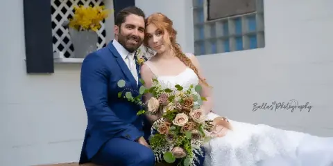 A bride and groom posing for a wedding portrait in front of a white building with a wooden bench.