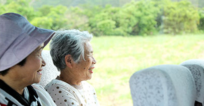 Two smiling passengers seated inside a bus looking out the window at a green landscape.