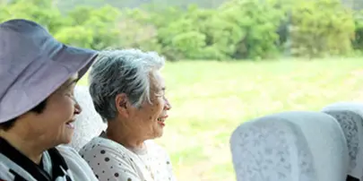 Two smiling passengers seated inside a bus looking out the window at a green landscape.