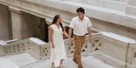 A couple holding hands and walking down a marble staircase in a grand building.