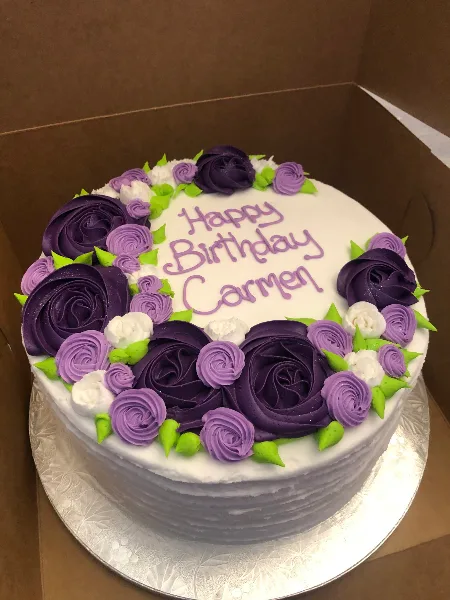 A round white cake decorated with purple and white piped frosting roses and the text 'Happy Birthday Carmen'.