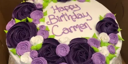 A round white cake decorated with purple and white piped frosting roses and the text 'Happy Birthday Carmen'.
