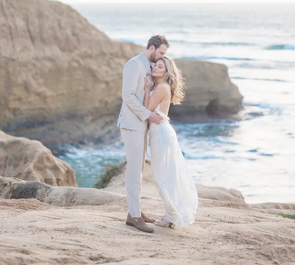 A bride and groom embracing on a rocky coastal cliff overlooking the ocean.