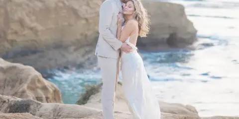 A bride and groom embracing on a rocky coastal cliff overlooking the ocean.