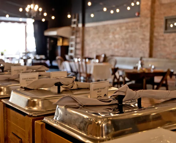 A catering buffet line featuring stainless steel chafing dishes with food labels in a rustic event venue.