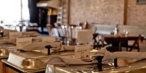 A catering buffet line featuring stainless steel chafing dishes with food labels in a rustic event venue.