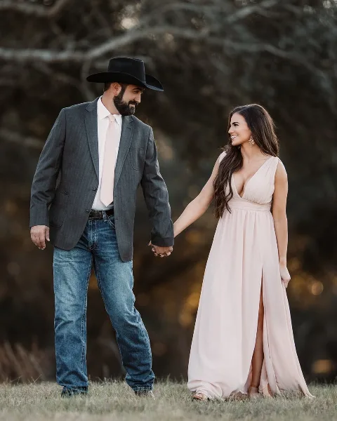 A professional engagement photograph of a couple holding hands in a field; the man wears a grey suit and cowboy hat, and the woman wears a long pink dress.