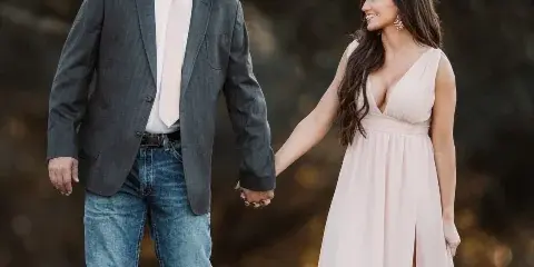 A professional engagement photograph of a couple holding hands in a field; the man wears a grey suit and cowboy hat, and the woman wears a long pink dress.
