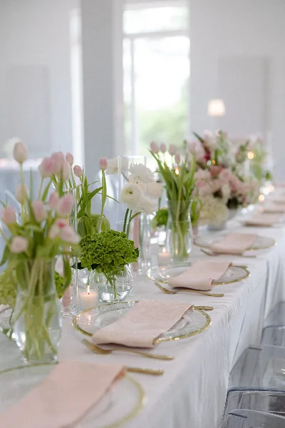 A long banquet table set with white linens, gold-rimmed glassware, pink napkins, and tall floral centerpieces featuring pink tulips and white ranunculus.