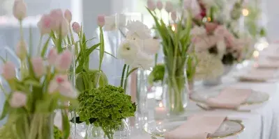 A long banquet table set with white linens, gold-rimmed glassware, pink napkins, and tall floral centerpieces featuring pink tulips and white ranunculus.