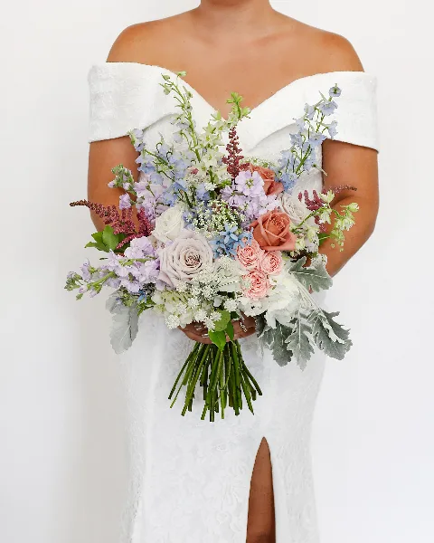 A bride in a white off-the-shoulder gown holding a large, mixed-color bridal bouquet featuring roses, delphiniums, and greenery.
