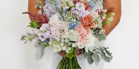 A bride in a white off-the-shoulder gown holding a large, mixed-color bridal bouquet featuring roses, delphiniums, and greenery.