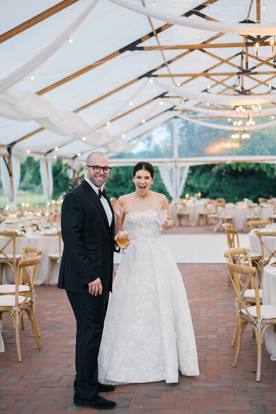 A bride and groom standing in a decorated wedding tent with string lights and set tables.