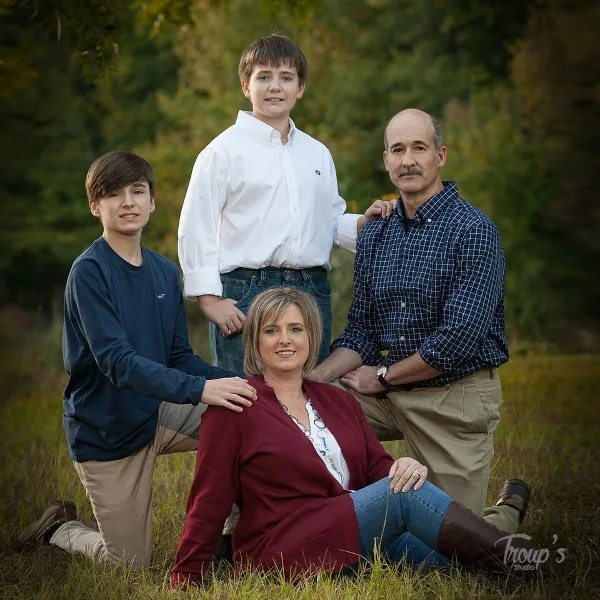 A professional outdoor family portrait of four people sitting and standing in a grassy field, watermarked with Troup's Studio.