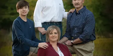 A professional outdoor family portrait of four people sitting and standing in a grassy field, watermarked with Troup's Studio.