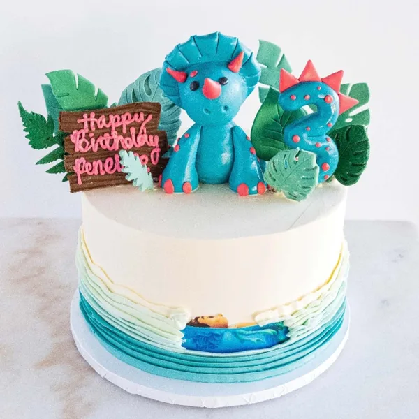 A white frosted cake decorated with a blue fondant dinosaur, green leaves, and a number 3 candle.