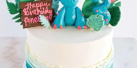 A white frosted cake decorated with a blue fondant dinosaur, green leaves, and a number 3 candle.